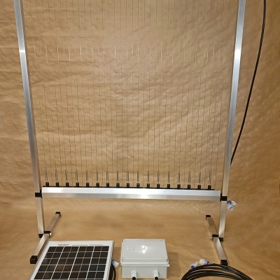 Estrutura metálica com fios verticais, painel solar, caixa elétrica e cabos em fundo castanho.