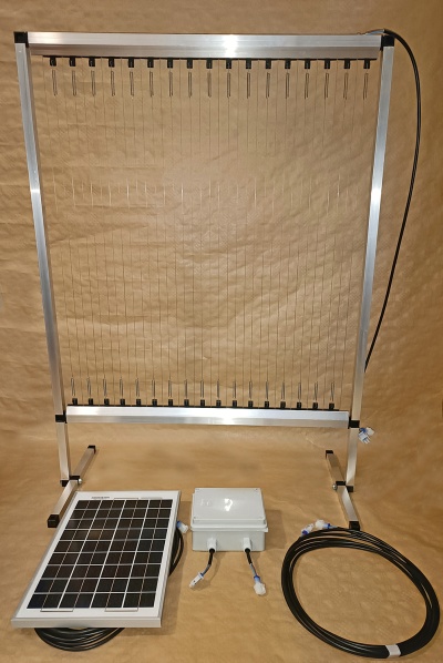 Estrutura metálica com fios verticais, painel solar, caixa elétrica e cabos em fundo castanho.