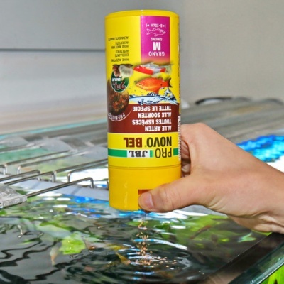 Frasco amarelo de alimento para peixes PRO JBL NOVO BEL segurado sobre aquário com peixes coloridos