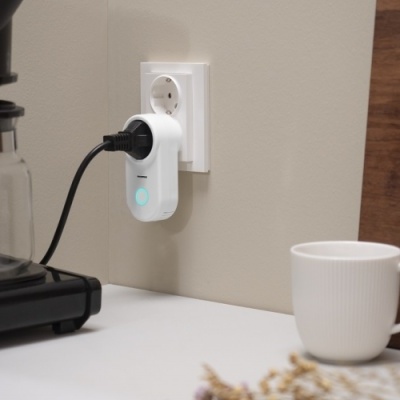 Cafeteira preta com jarro de vidro e adaptador branco ligado a tomada na parede ao lado de uma caneca branca