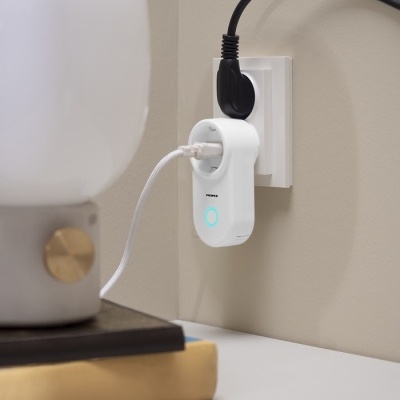 Ficha elétrica branca com cabo USB ligada em tomada na parede junto a lâmpada de mesa e livro