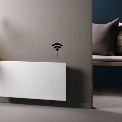 Radiador elétrico branco com símbolo Wi-Fi na parede cinza ao lado de mesa com planta e banco com almofadas.