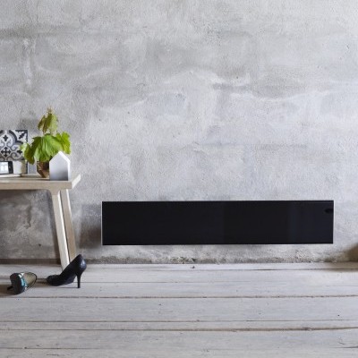 Radiador preto montado na parede cinzenta junto a banco de madeira clara com planta e sapatos no chão