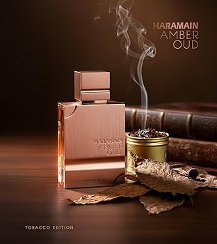 Frasco de perfume Haramain Amber Oud com folhas de tabaco e livro antigo em fundo escuro