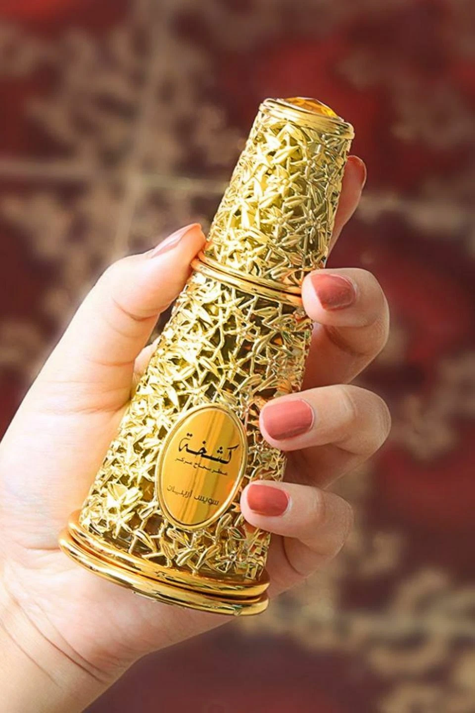 Frasco de perfume dourado intrincado com rótulo em árabe, segurado por uma mão com unhas coral.