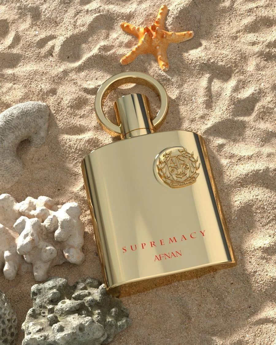 Frasco de perfume dourado SUPREMACY AFNAN na areia com estrela-do-mar e corais