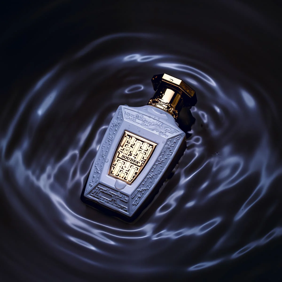 Frasco de perfume cinzento com detalhes dourados num líquido escuro ondulado