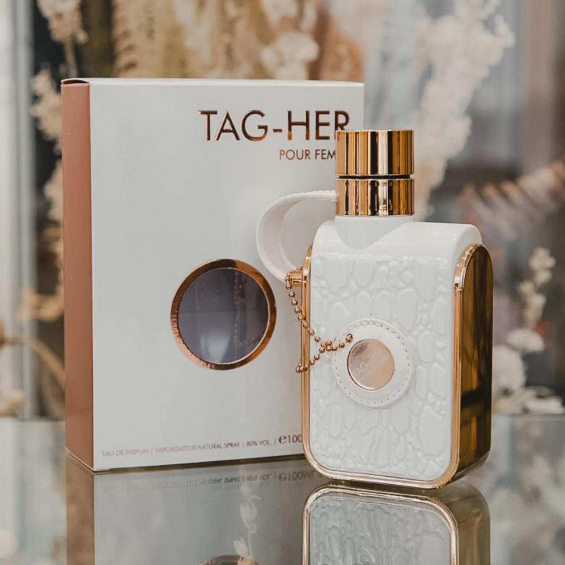 Frasco de perfume TAG-HER POUR FEMME branco e dourado ao lado da caixa correspondente.