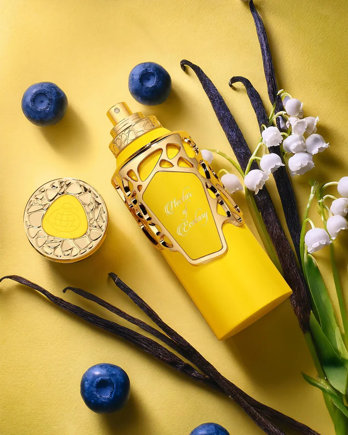 Frasco amarelo de perfume com tampa dourada, bagas azuis, vagens de baunilha e flores brancas num fundo amarelo