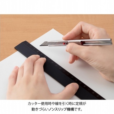 Mãos a usar régua preta e faca de precisão sobre papel branco numa mesa de madeira