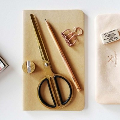 Material de papelaria com caderno bege, tesoura dourada, lápis, apontador, borracha e estojo de couro claro.