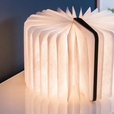 Lâmpada livro aberta iluminada sobre móvel branco com caderno e planta ao fundo