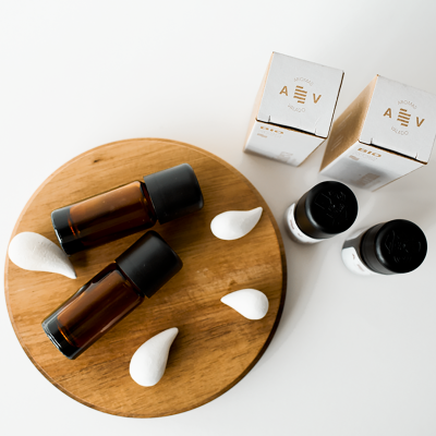 Frascos de vidro castanho com tampas pretas sobre tábua de madeira redonda, formas brancas, embalagens brancas com texto dourado e frascos pequenos