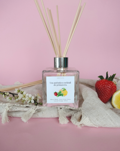 Difusor Mikado em vidro com aromatizador e varas de madeira sobre tecido bege, com morango, limão e flores, fundo rosa.