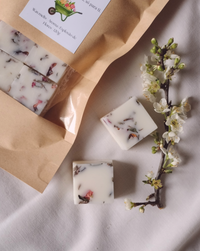 Blocos quadrados de cera aromática com pétalas, saco de papel castanho e ramo florido em tecido branco.