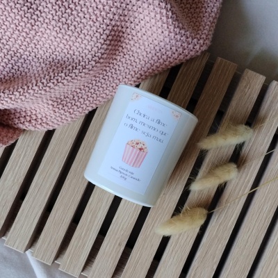 Vela aromática branca com etiqueta e manta rosa tricotada sobre banco de madeira claro.