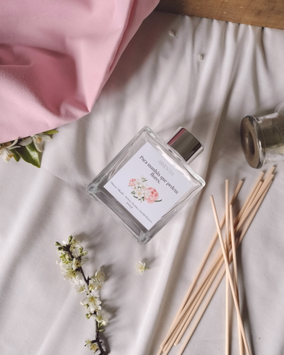 Frasco de vidro quadrado com tampa prateada e palitos de difusão de madeira, com fundo branco e tecido rosa.