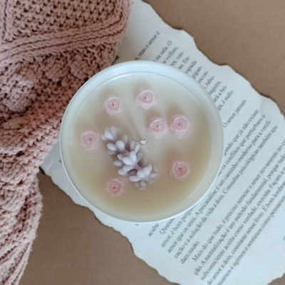 Vela branca com flores decorativas rosas e lilás sobre página de livro e tricot rosa.