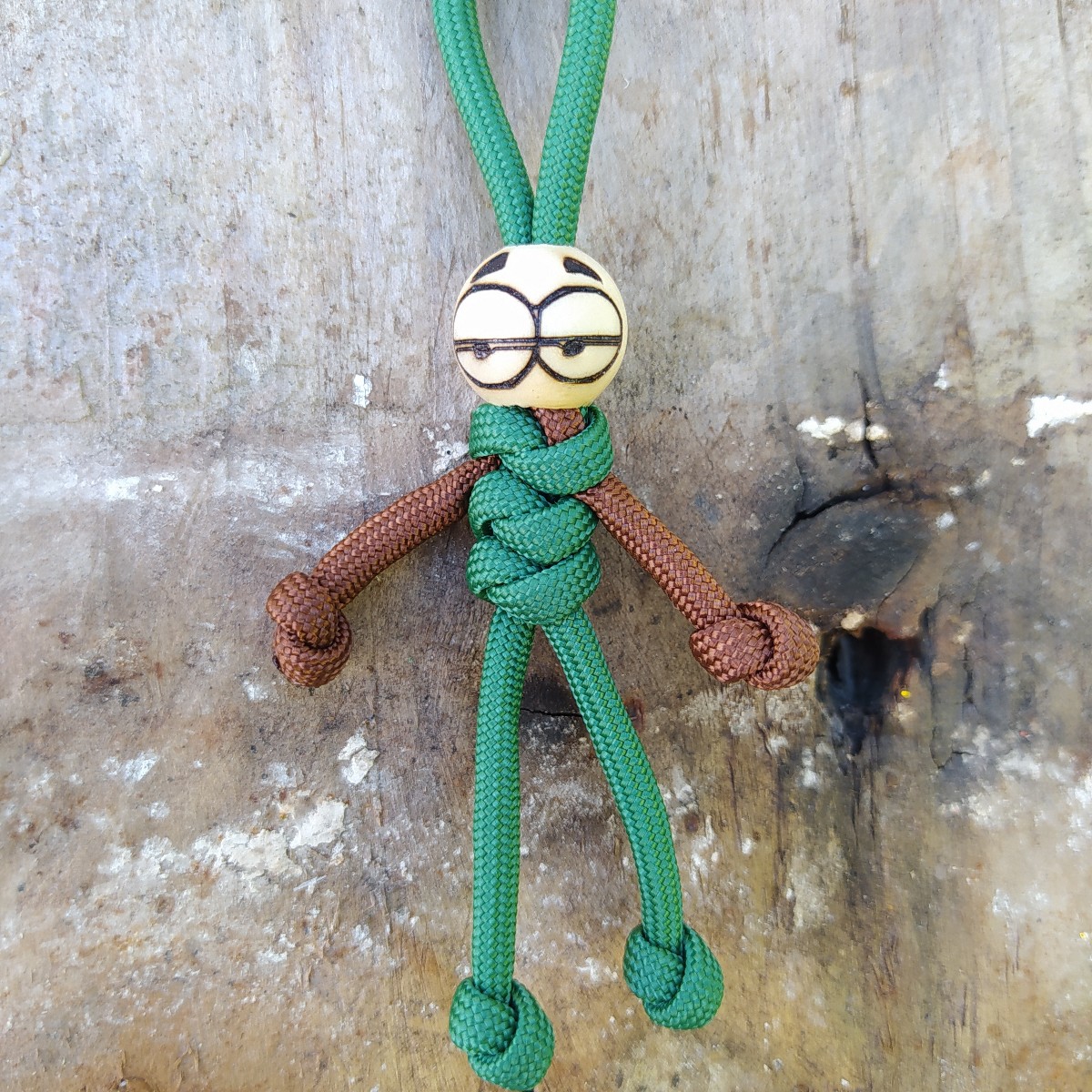 Boneco feito de cordas verde e castanha com cabeça branca desenhada, pendurado num fundo de madeira