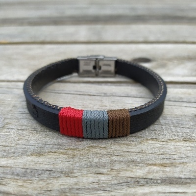 Pulseira em couro preto com fios trançados vermelho, cinza e castanho e fecho metálico sobre madeira.