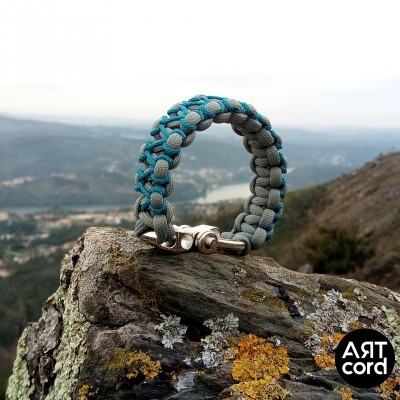 Pulseira trançada de tecido azul e cinzento com fecho metálico sobre rocha com líquenes
