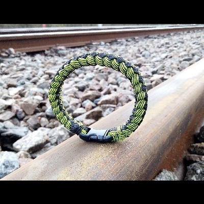 Pulseira trançada preta e verde com fecho preto sobre carril de comboio.