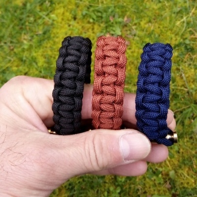 Três pulseiras de corda trançada preta, castanha e azul escuro na mão sobre relva verde.