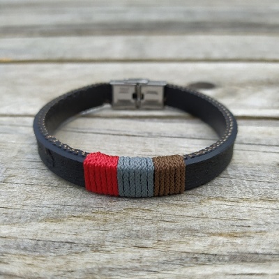 Pulseira em couro preto com fios trançados vermelho, cinza e castanho e fecho metálico sobre madeira.