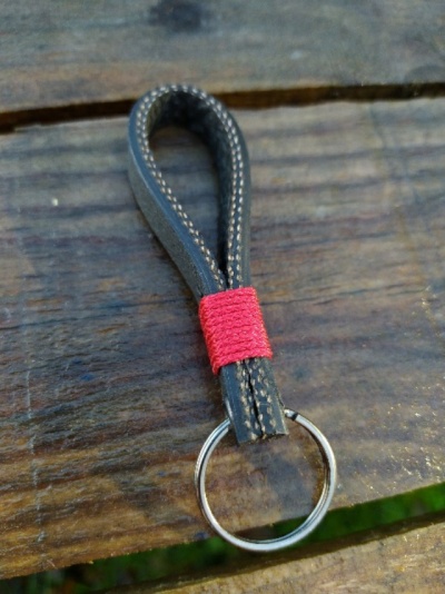 Porta-chaves em fita preta com costura branca e fio vermelho, anel metálico, sobre madeira escura