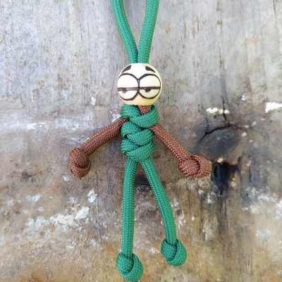 Boneco feito de cordas verde e castanha com cabeça branca desenhada, pendurado num fundo de madeira