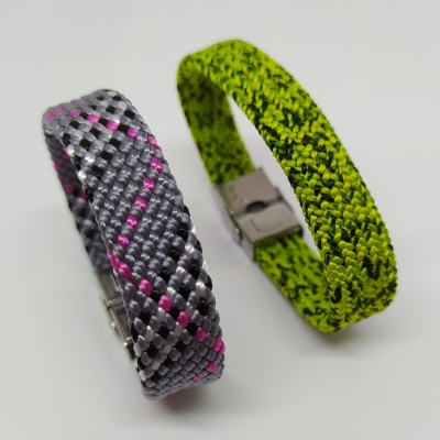 Duas pulseiras trançadas de tecido, uma cinzenta com rosa e preta, outra verde com preto, com fecho metálico.