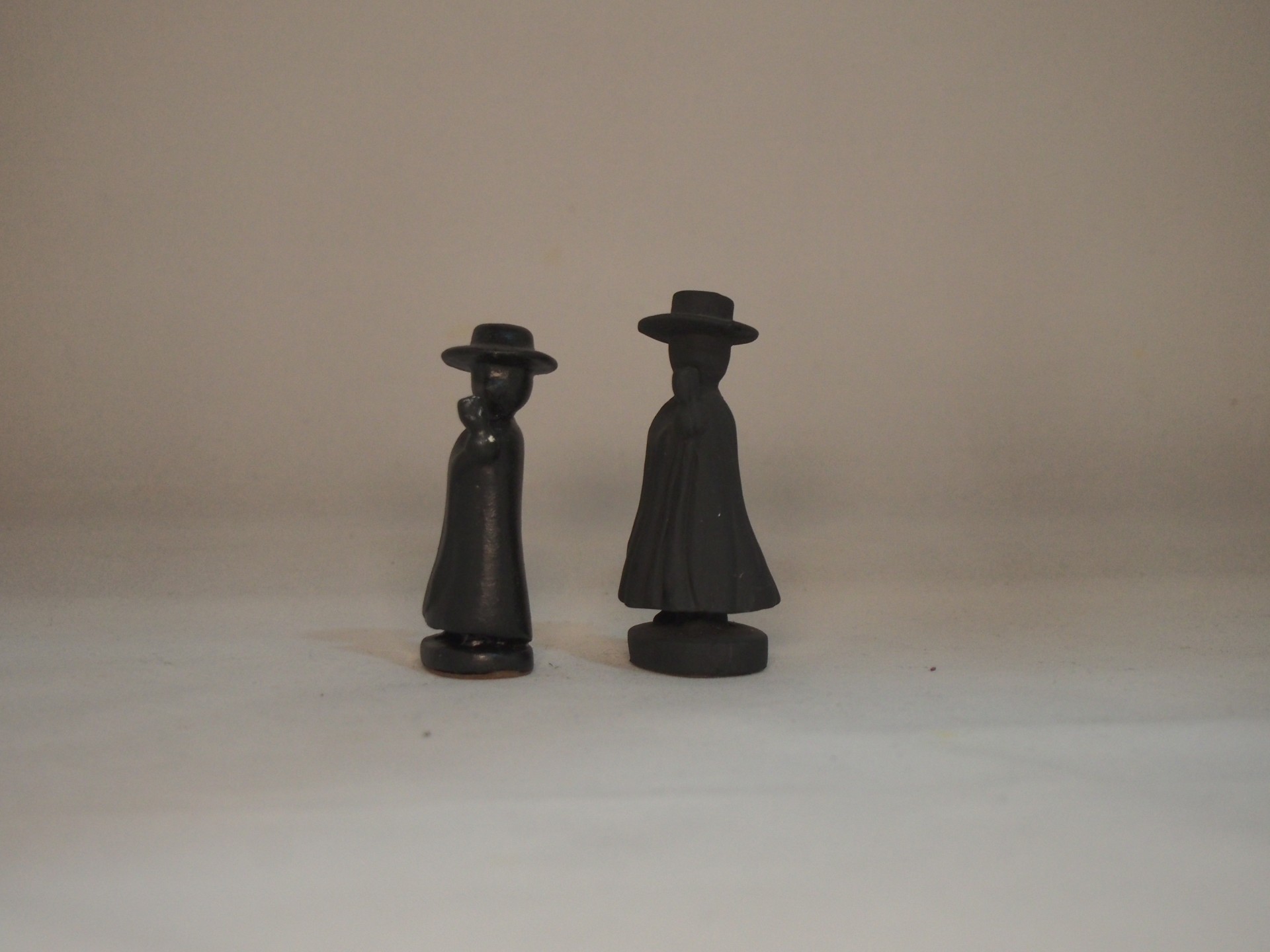Duas estatuetas pequenas pretas com chapéus e roupas compridas sobre superfície branca