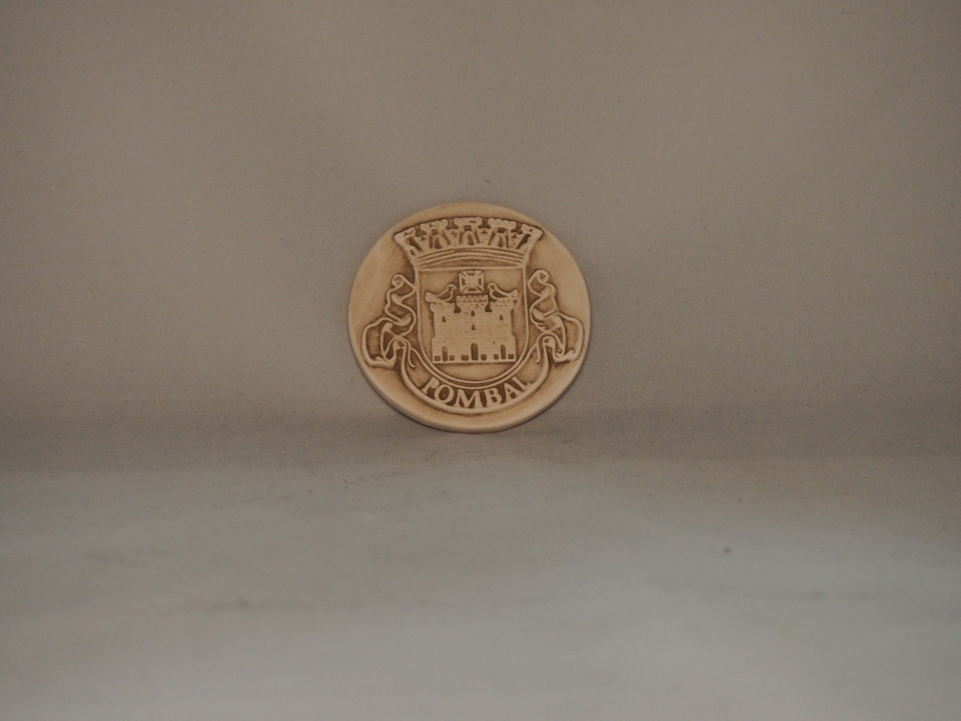 Moeda ou medalha bege com brasão e texto LOMBA sobre superfície cinza.