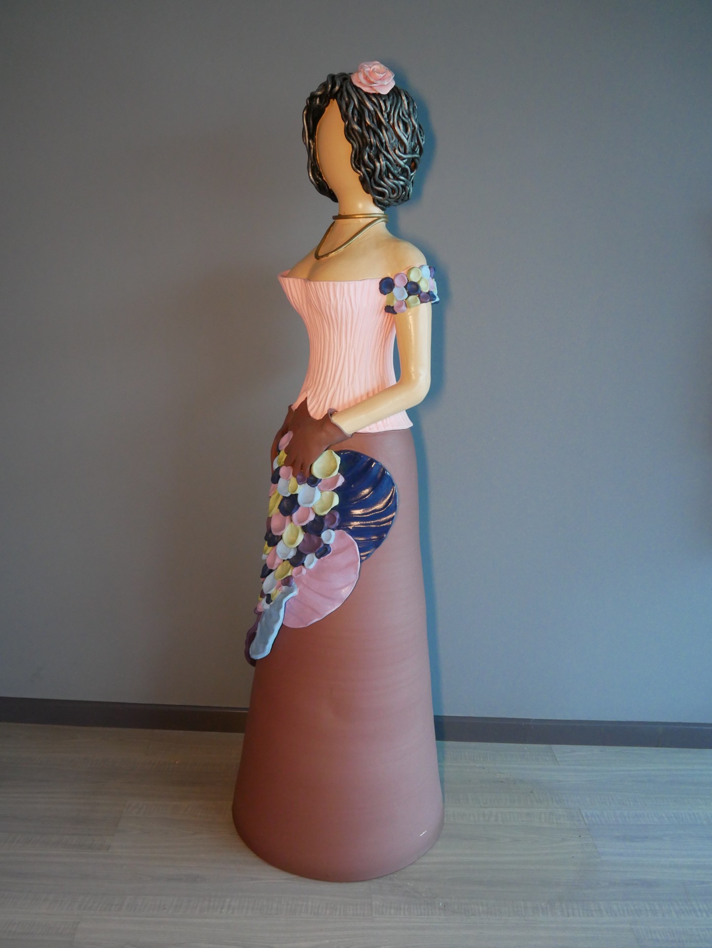 Estátua decorativa de mulher com vestido longo rosa e castanho, cabelo cacheado negro e flor rosa na cabeça.