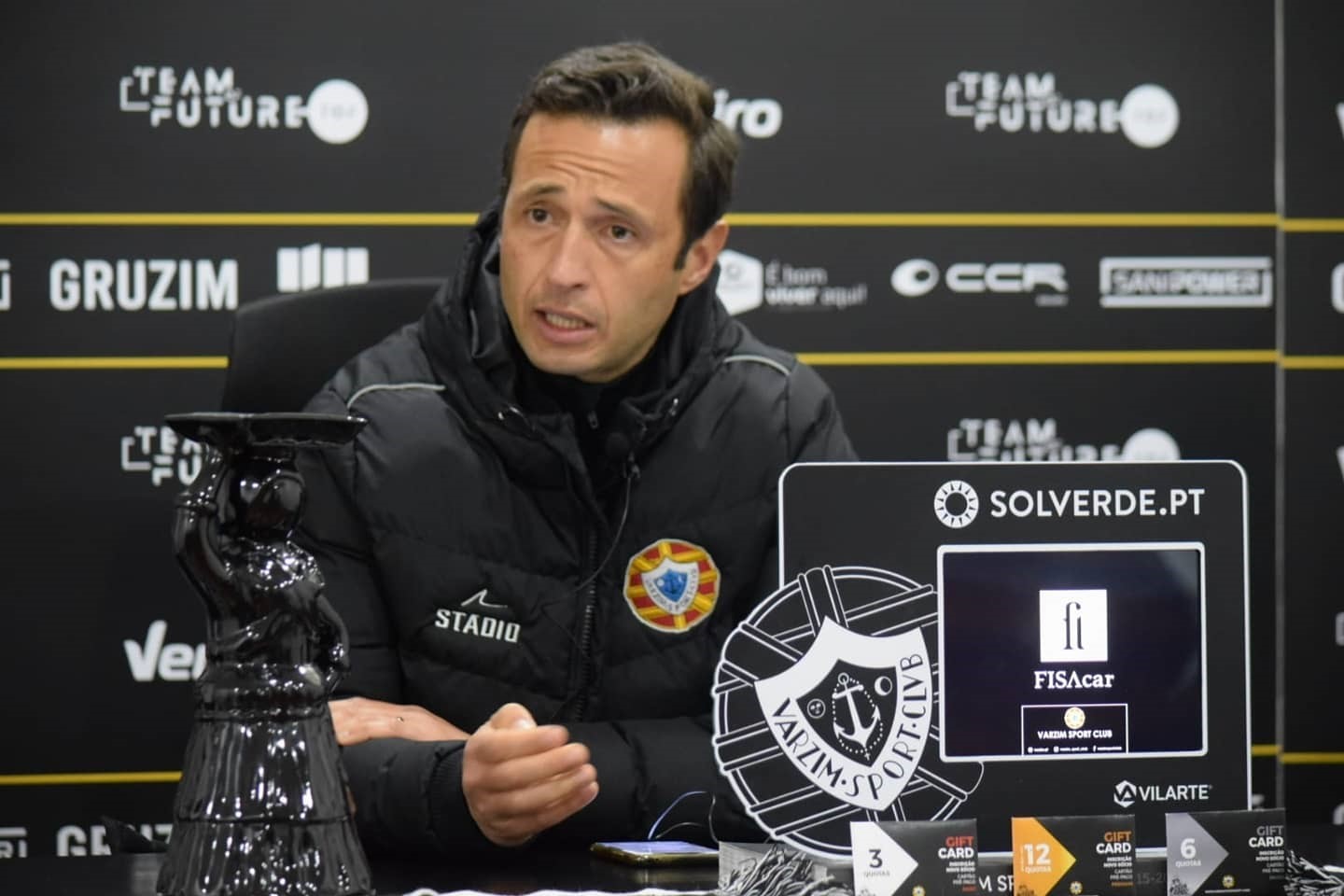 Homem em casaco preto em conferência com logótipos de marcas e clube Valim-Sport em fundo escuro.