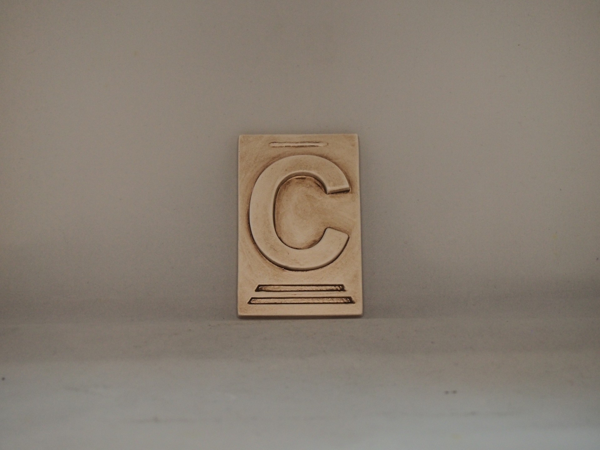 Placa decorativa bege com a letra 'C' em relevo