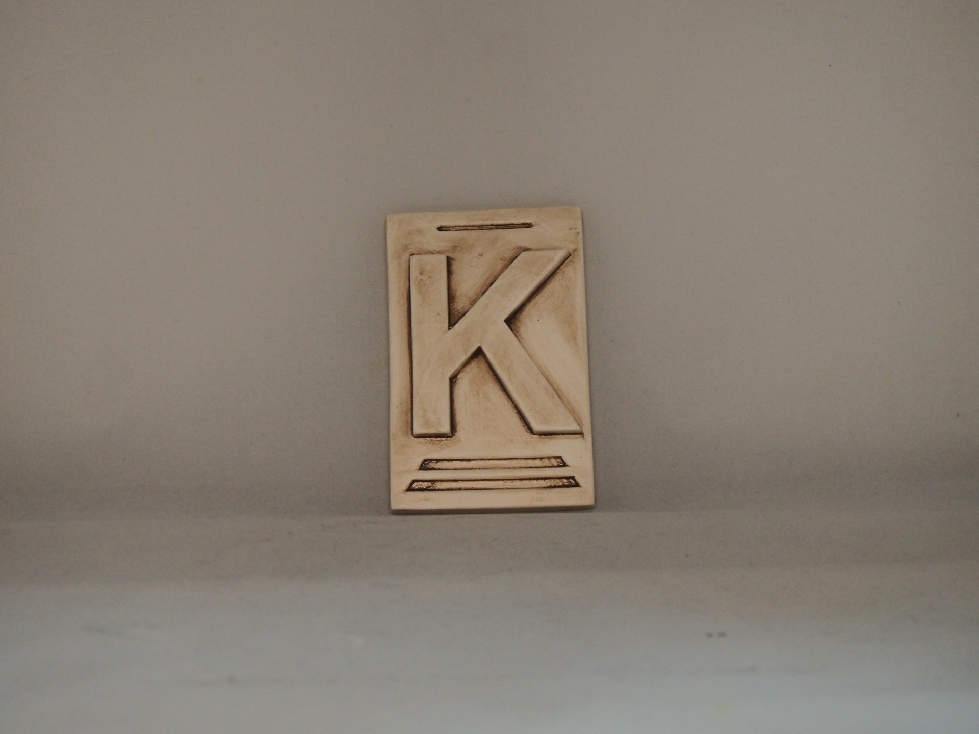 Placa decorativa bege com letra K em relevo