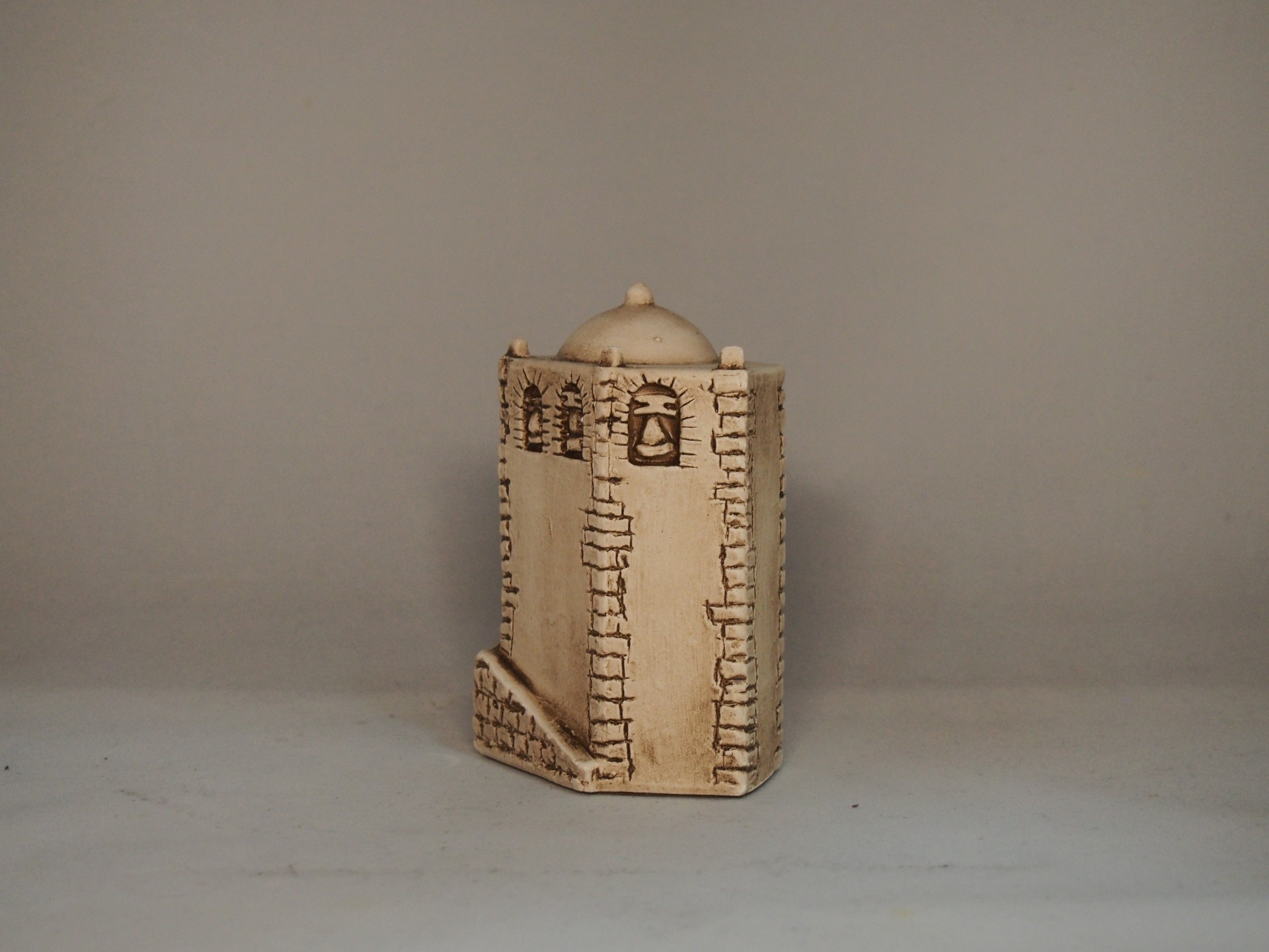 Escultura cerâmica castanho claro com padrão de tijolos e sinos, em fundo cinzento.