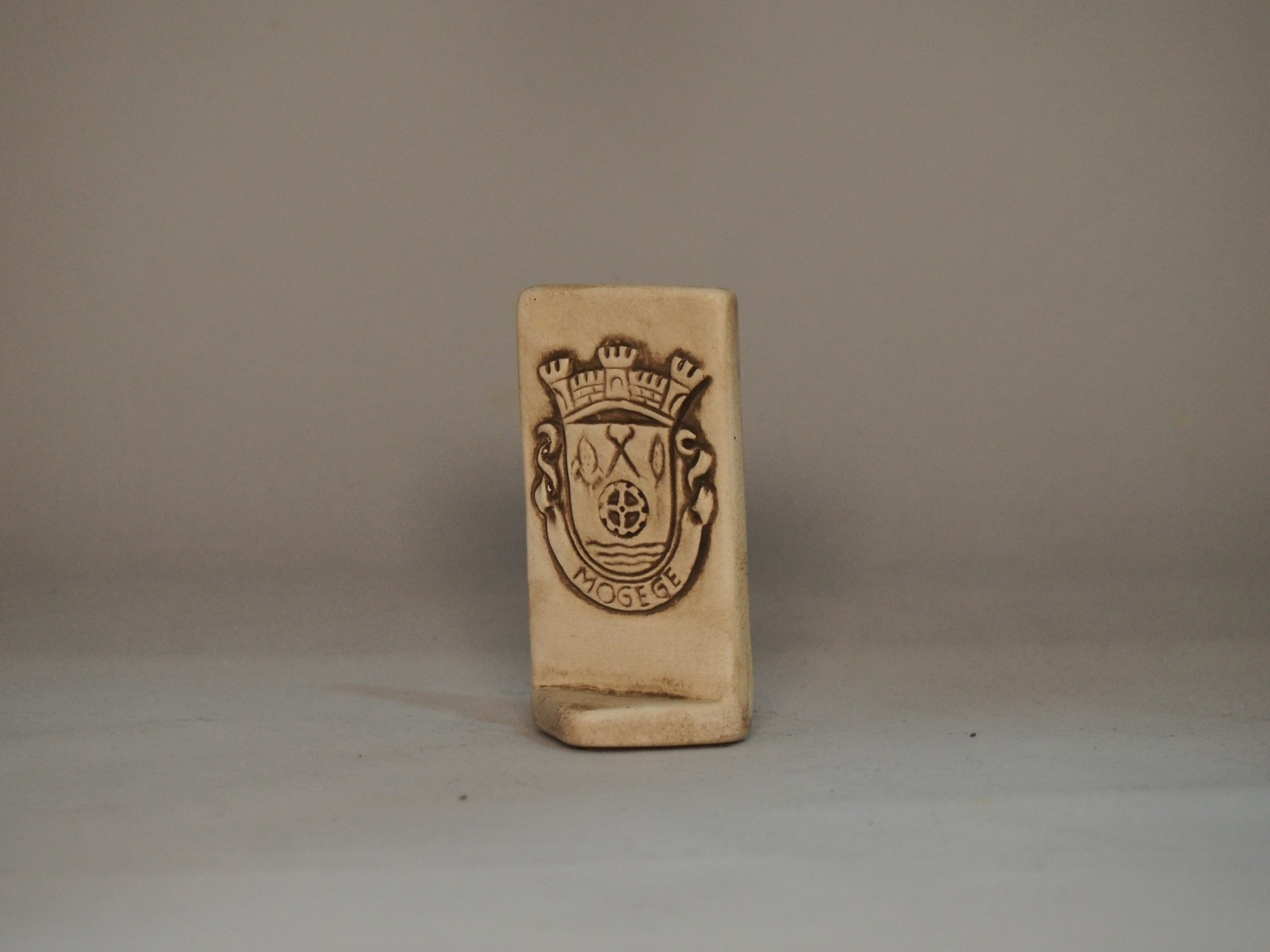 Objeto de cerâmica bege com brasão e texto 'XO Moises' sobre fundo cinzento