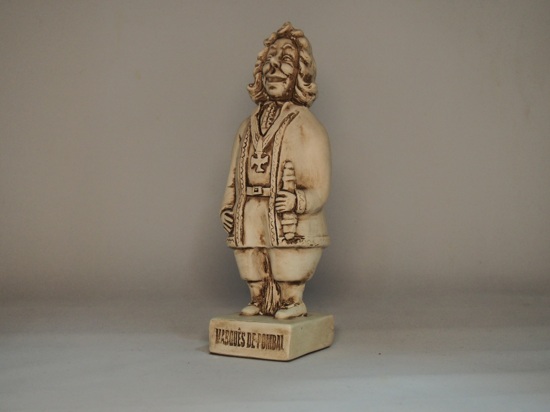 Estátua bege claro de figura masculina com túnica, segurando objeto, sobre base com texto