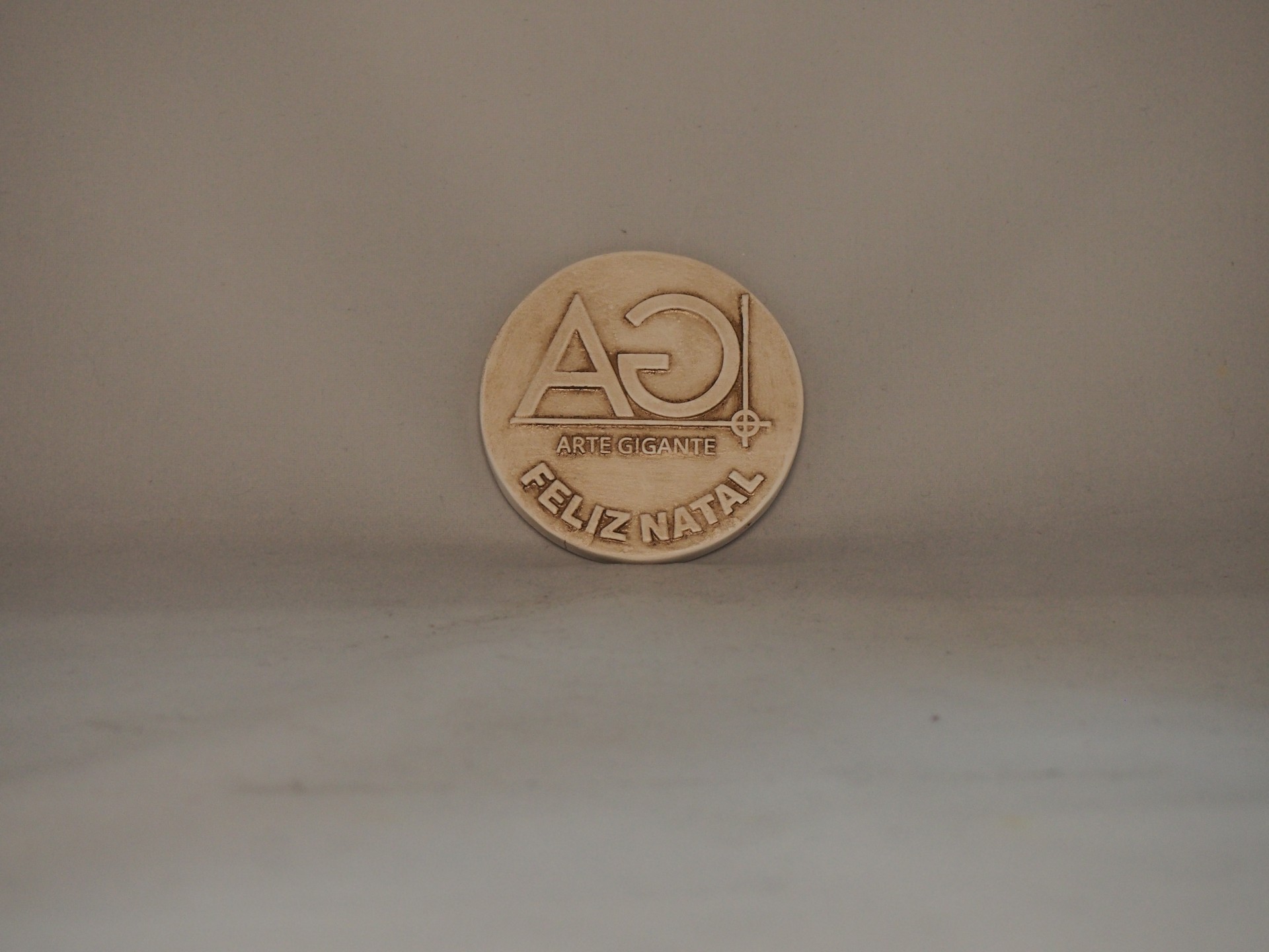 Moeda decorativa com letras AG e texto FELIZ NATAL em relevo