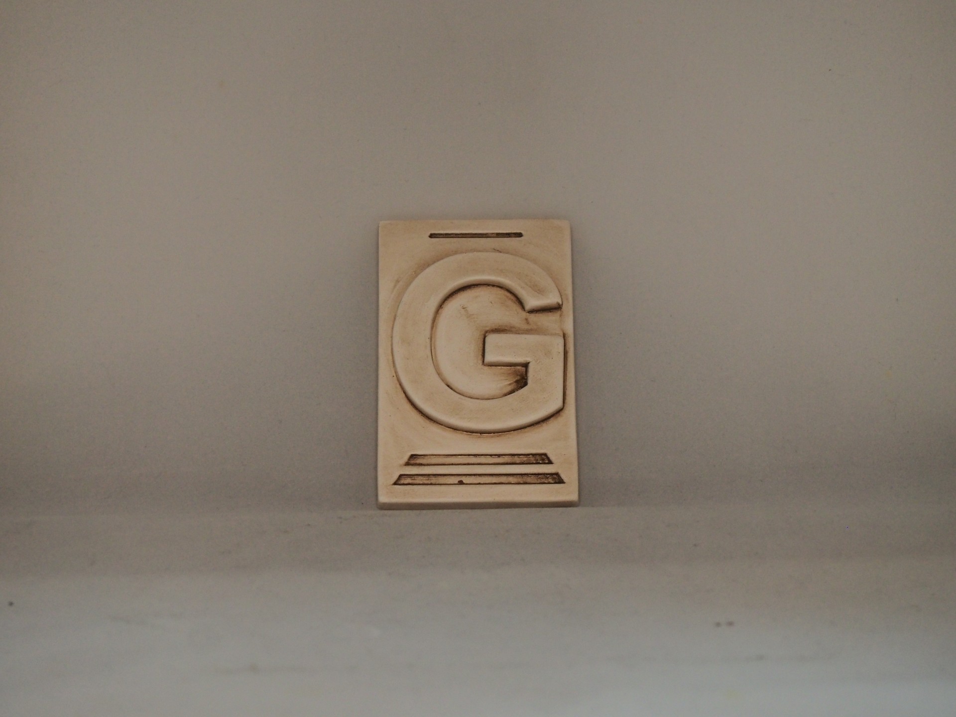 Peça decorativa bege com relevo da letra G