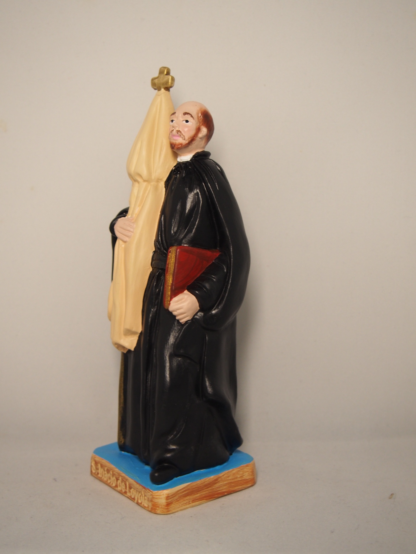 Figurinha de frade com hábito preto, segurando livro vermelho e bandeira bege com cruz dourada