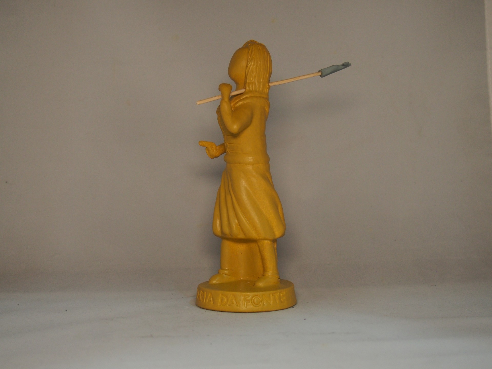 Estátua amarela de guerreiro indígena com lança sobre fundo neutro