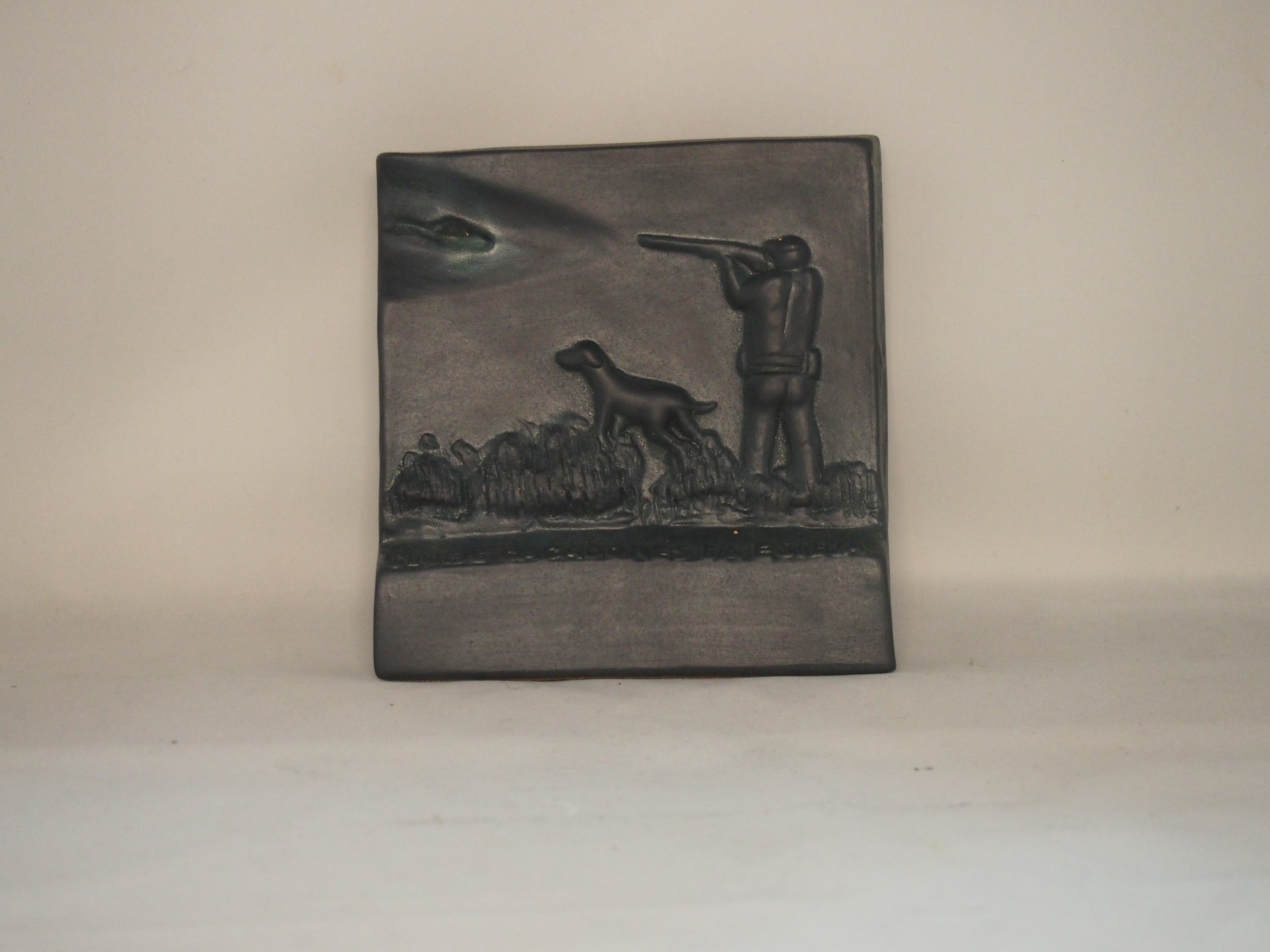 Placa decorativa em metal escuro com relevo de caçador e cão.