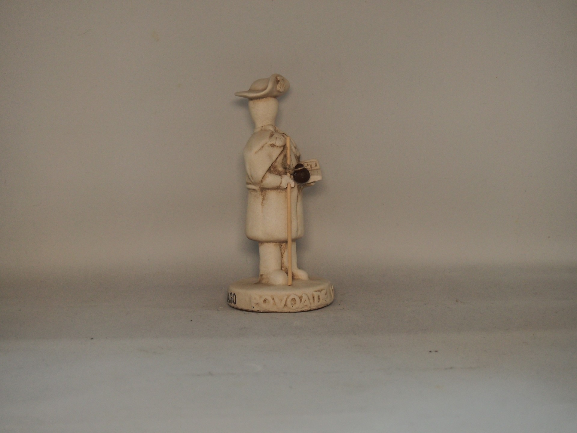 Estatueta pequena de figura humana em sobretudo e chapéu sobre base com texto