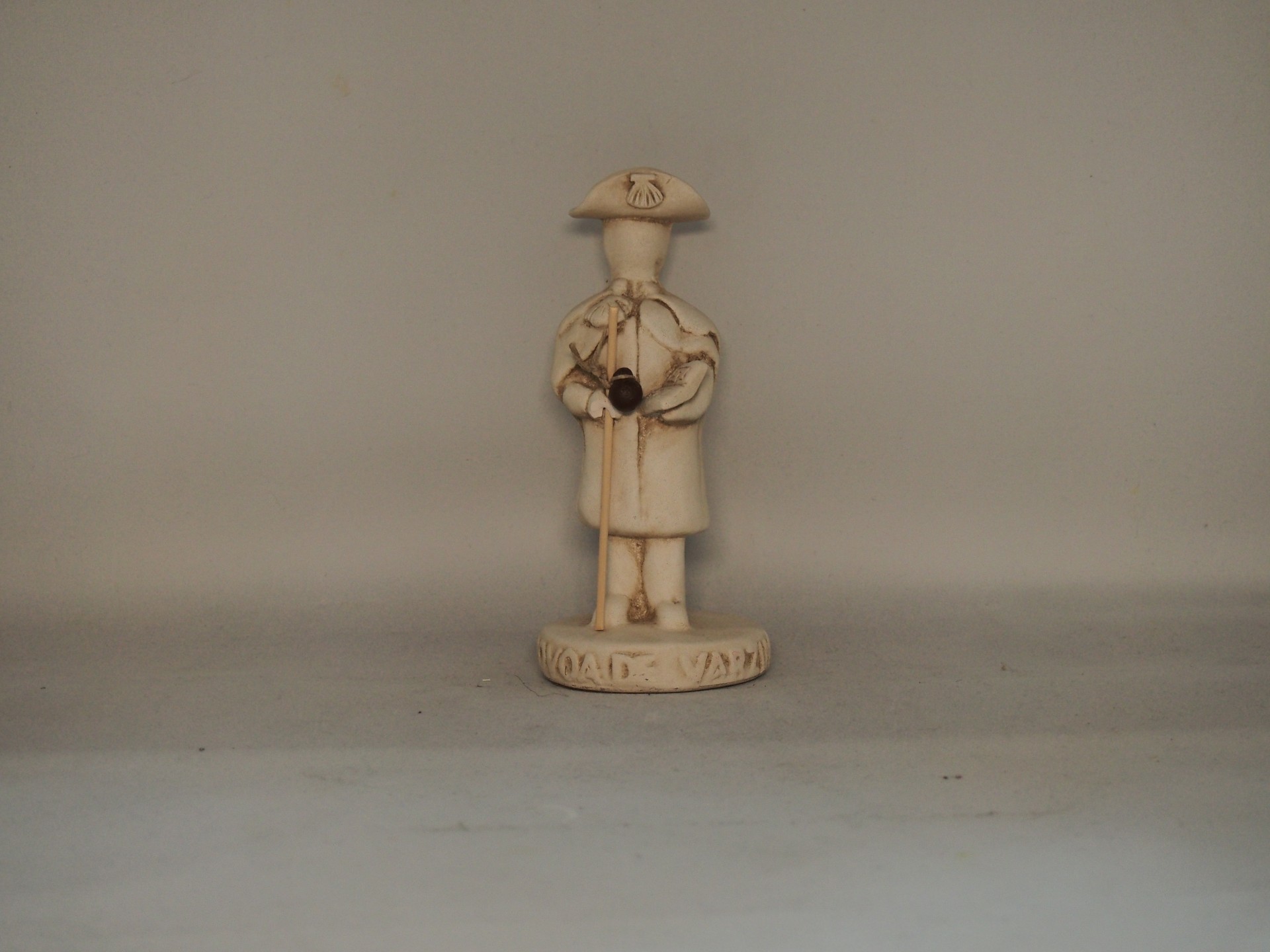 Figura decorativa bege com chapéu e bengala sobre base redonda em fundo cinzento.