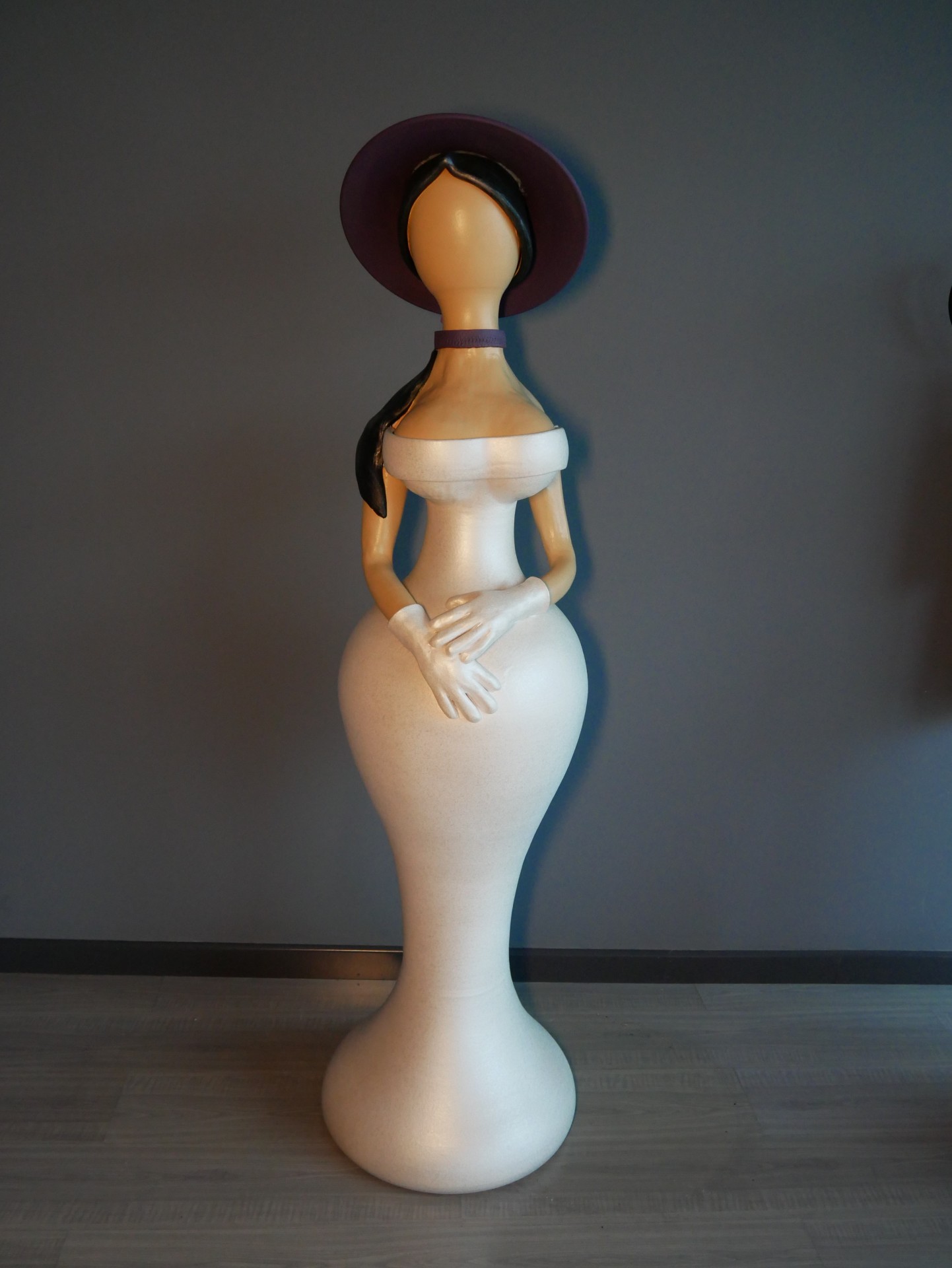 Estátua decorativa de mulher estilizada, vestido creme, chapéu roxo, em fundo cinza