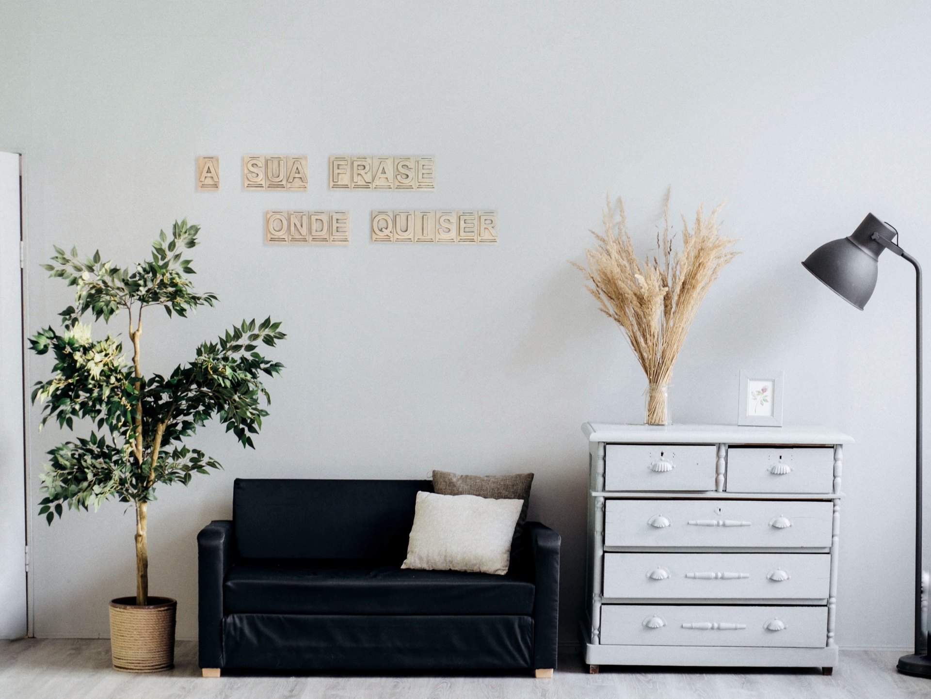 Sofá preto, cómoda branca, planta em vaso, candeeiro, parede com letras