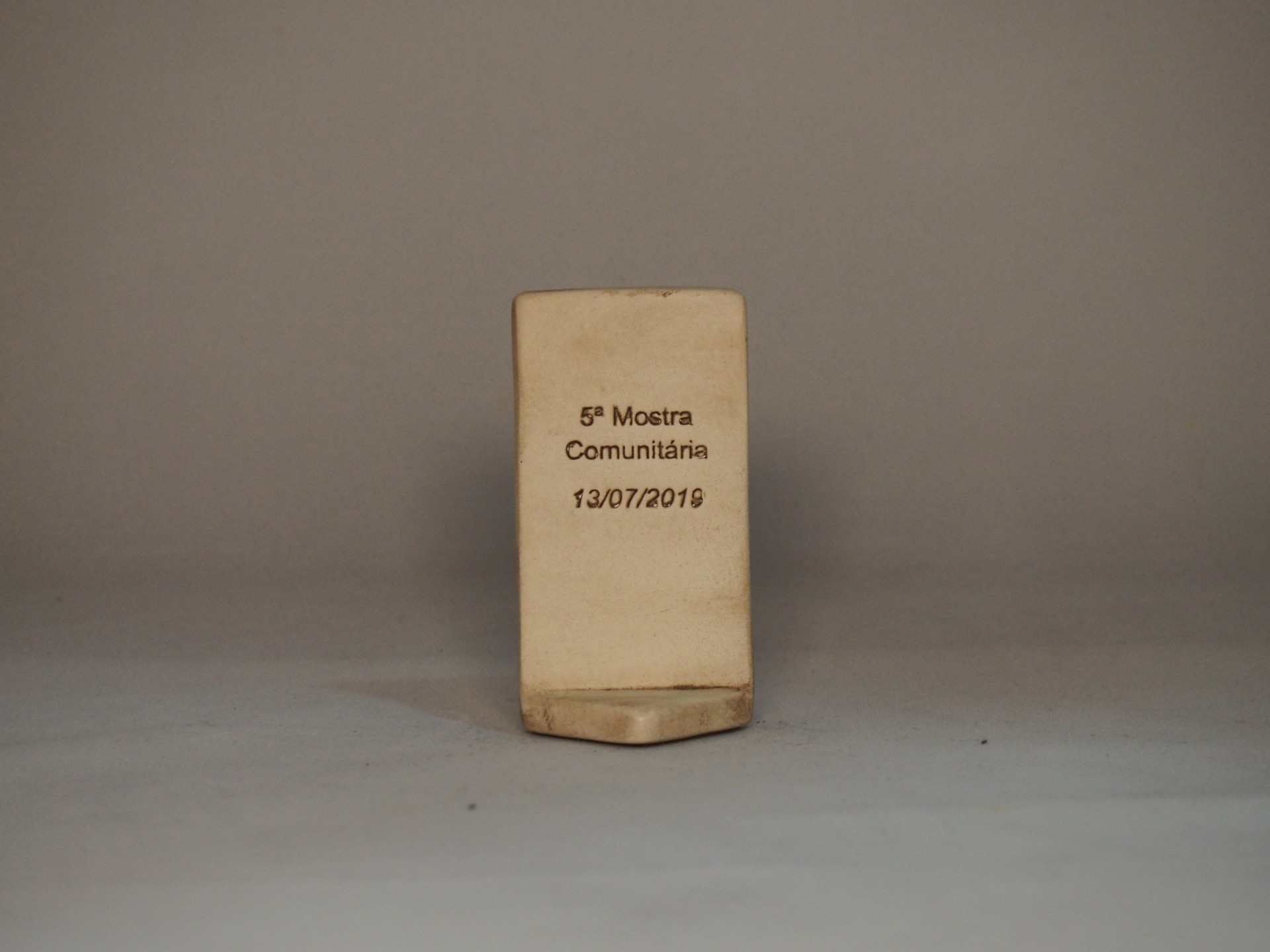 Placa ou troféu bege com inscrição 5ª Mostra Comunitária 13/07/2019